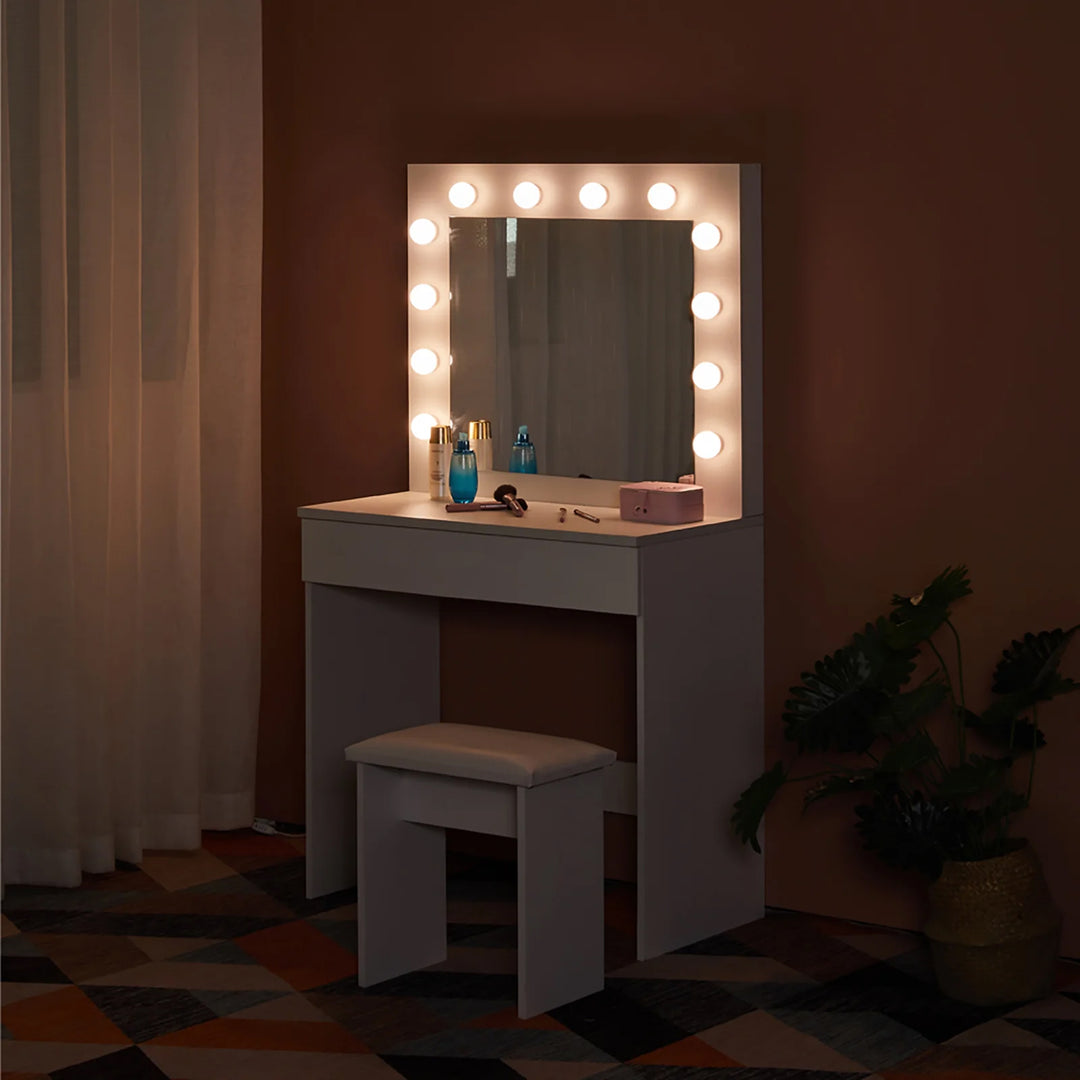 The side view of the white dressing table set, featuring a mirror, 12 LED bulbs, 1 drawer, and a stool, in a dark bedroom, with lights on.