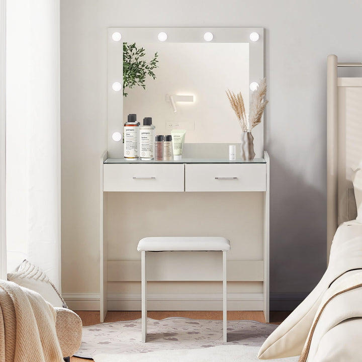 The front view of the white dressing table set, featuring a mirror, 8 LED bulbs, a glass tabletop, 2 drawers, and a stool, in a bright bedroom.