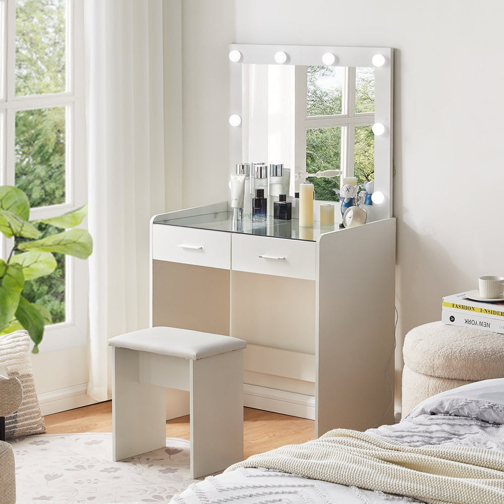 The side view of the white dressing table set, featuring a mirror, 8 LED bulbs, a glass tabletop, 2 drawers, and a stool, in a bright bedroom.