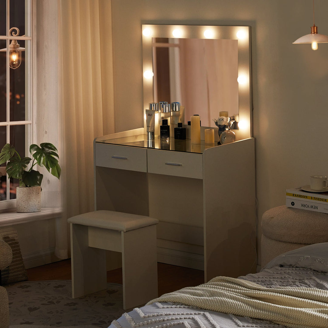 The side view of the white dressing table set, featuring a mirror, 8 LED bulbs, a glass tabletop, 2 drawers, and a stool, in a dark bedroom, with lights on.