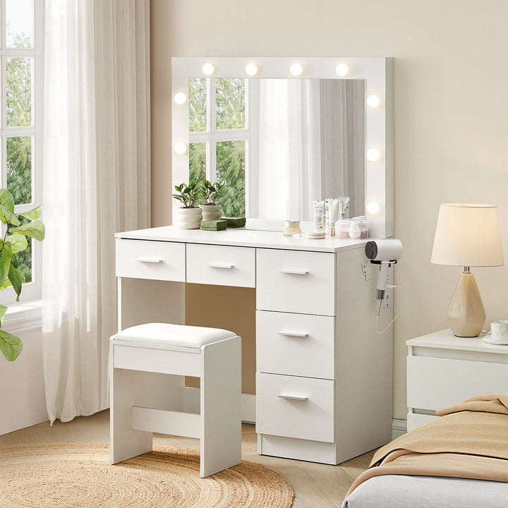 A side view of the 5-drawer Hollywood White Dressing Table with a stool, a large mirror, 10 bulbs in warm lighting mode, USB ports and power sockets, and a hairdryer holder in a bright bedroom