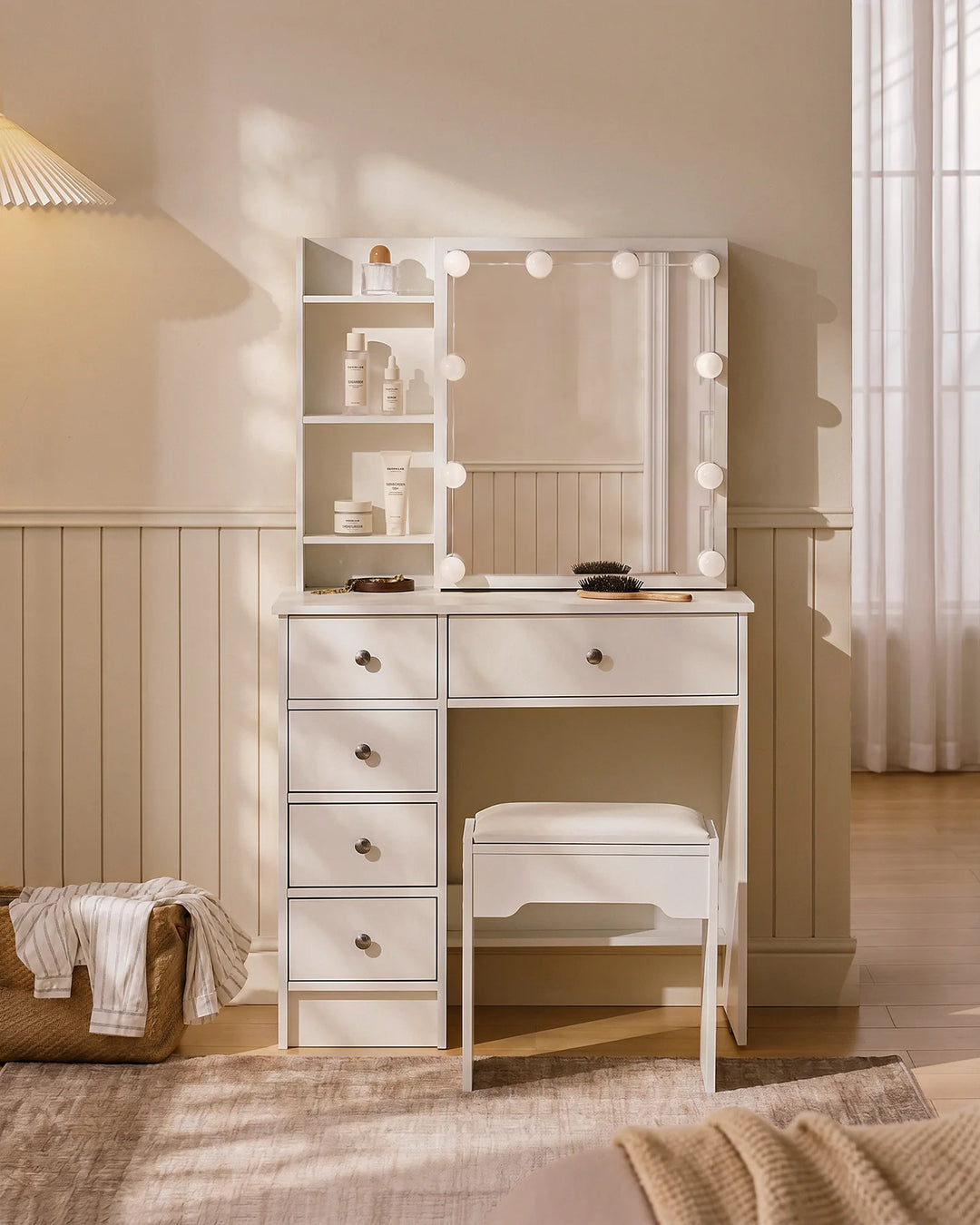 Front view of a white dressing table with bulbs, sliding mirror, hidden storage, open shelves, five drawers and a matching stool in a  small bedroom