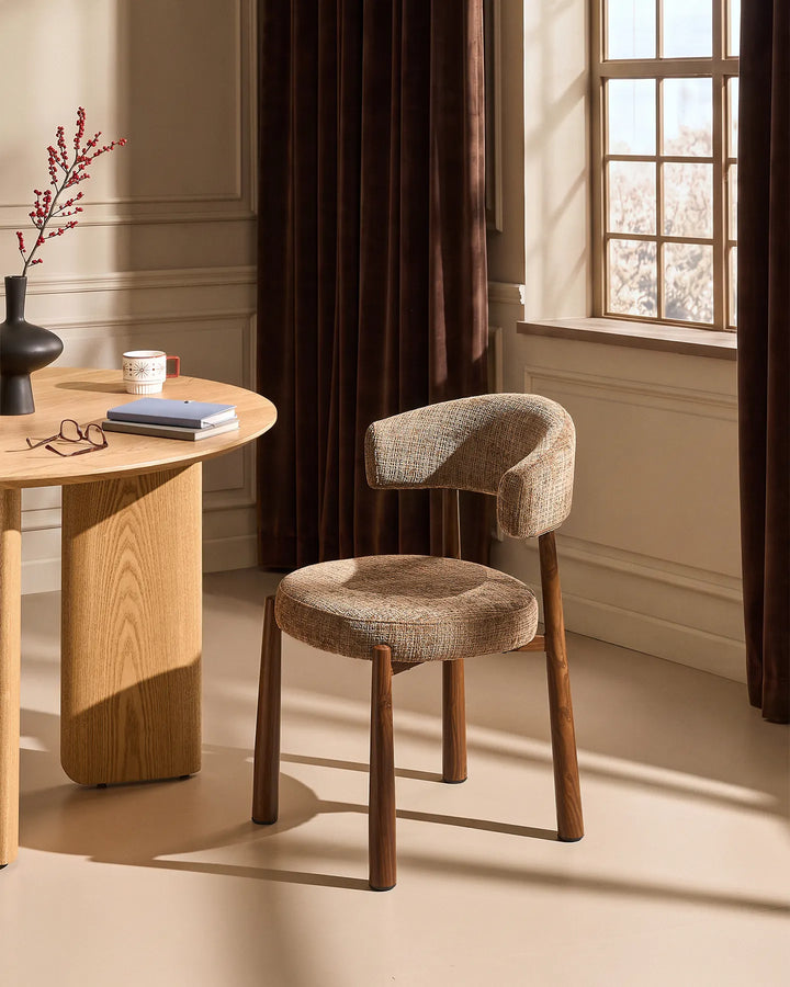 Ansel Mocha Chenille Dining Chair with curved upholstered backrest and wooden-effect legs beside a light oak dining table in a warm neutral dining room.