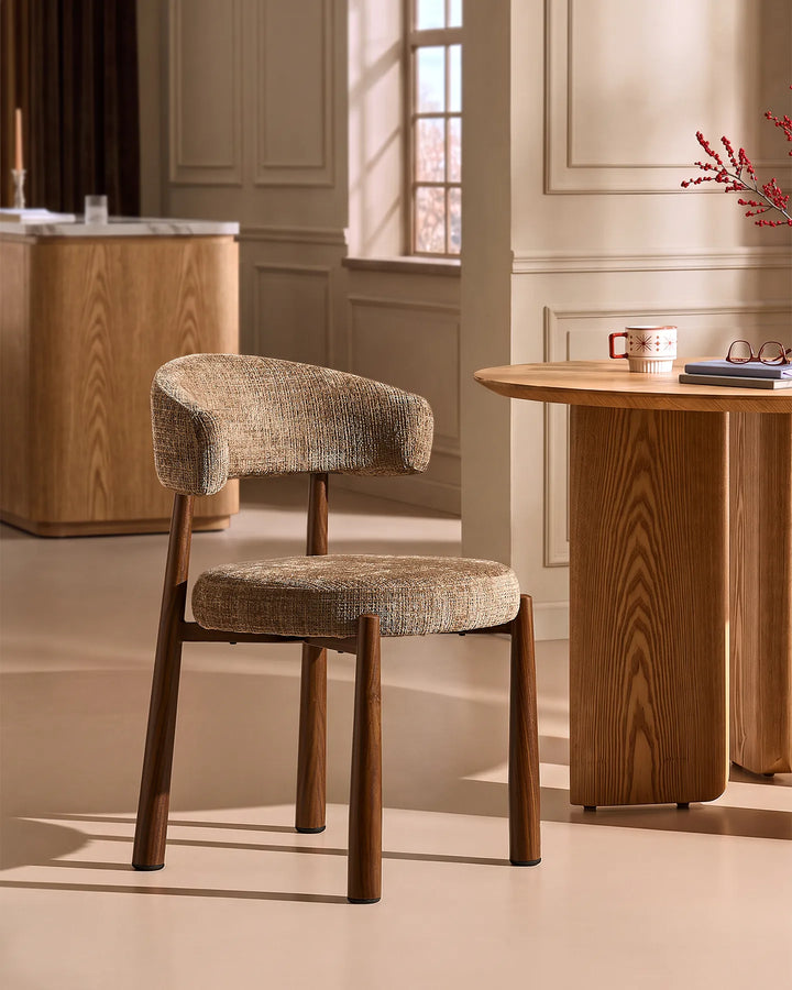 Side view of the Ansel Mocha Chenille Dining Chair beside a round oak dining table, highlighting its curved silhouette and compact design.