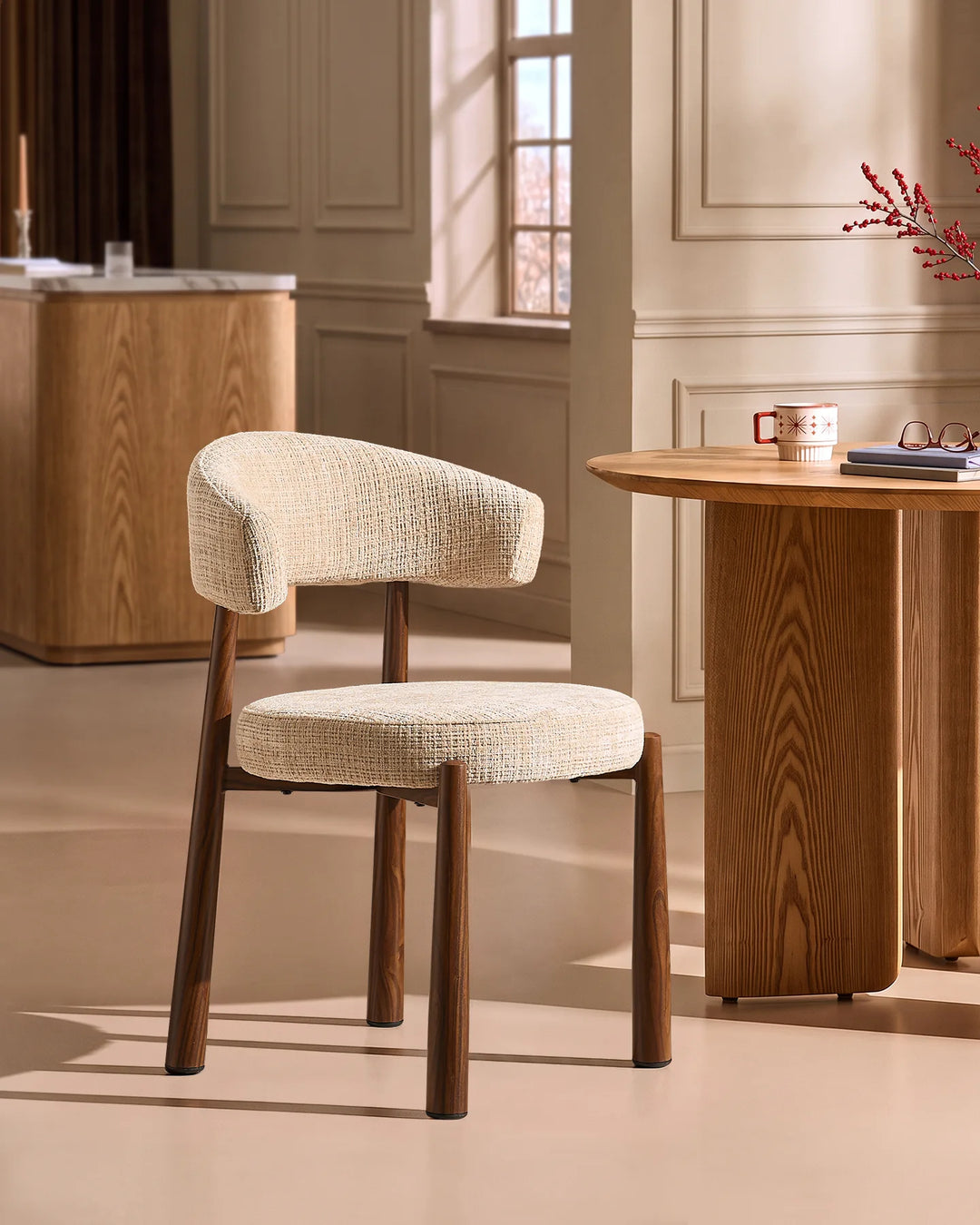 Side view of the Ansel Oatmeal Chenille Dining Chair beside a round oak dining table, highlighting its curved silhouette and compact design.