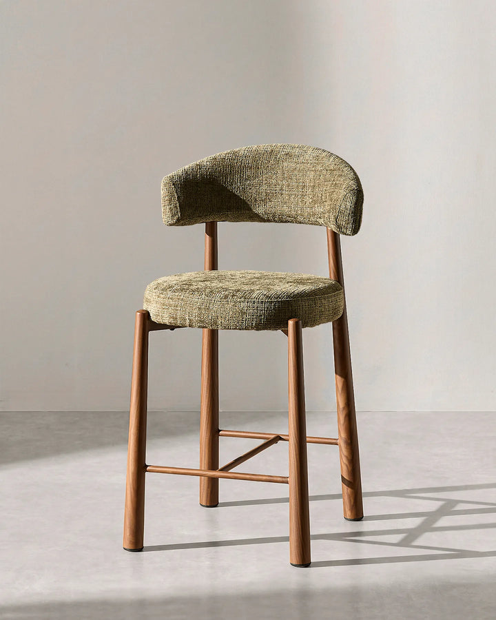 Front studio view of the Ansel bar stool highlighting the curved olive green chenille backrest, round cushioned seat, and wood-effect steel legs on a neutral background
