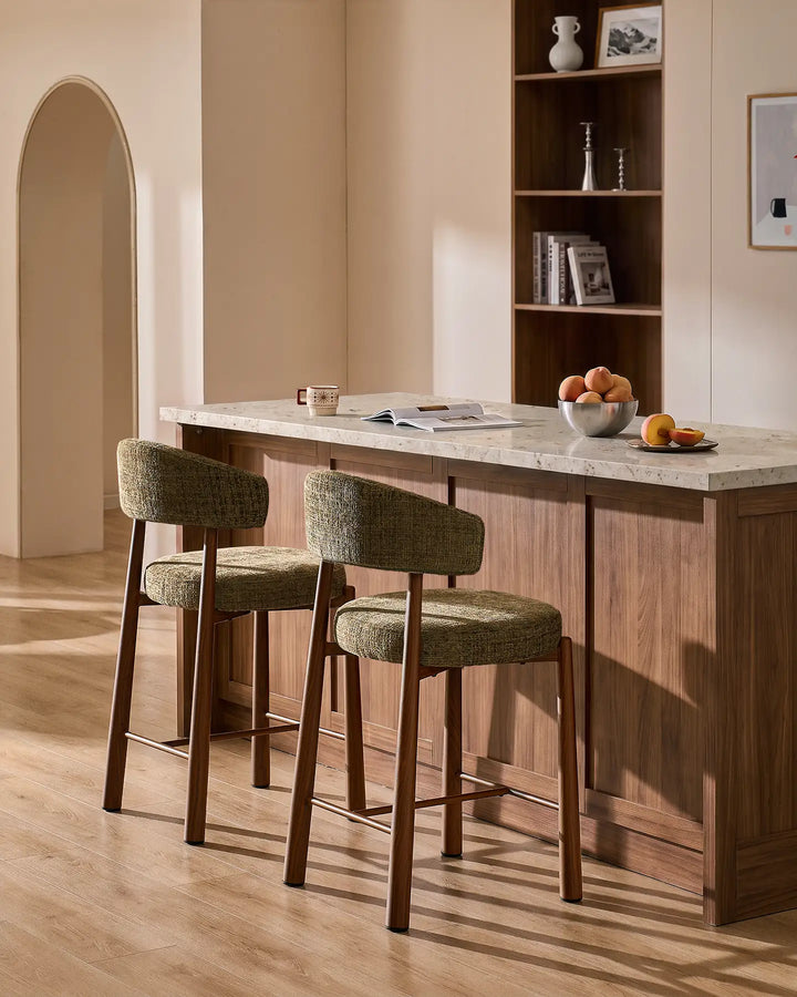 Two Ansel olive green chenille breakfast bar stools with wood-effect metal legs styled at a kitchen island with neutral terrazzo countertop and warm interior decor