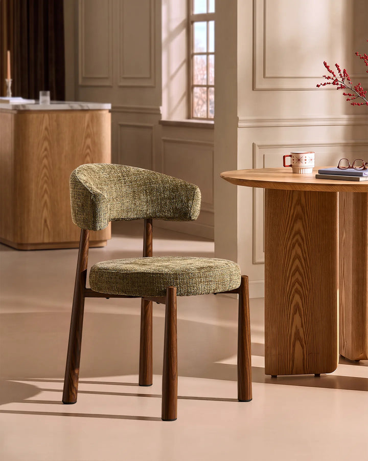 Side view of the Ansel Olive Green Chenille Dining Chair beside a round oak dining table, highlighting its curved silhouette and compact design.