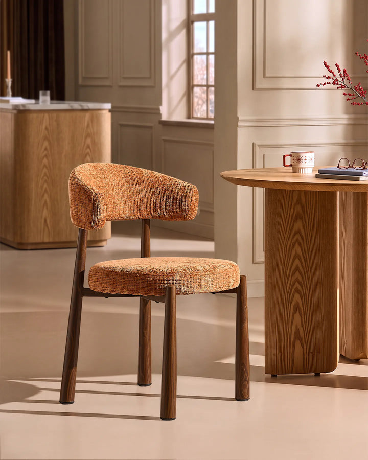 Side view of the Ansel Terracotta Chenille Dining Chair beside a round oak dining table, highlighting its curved silhouette and compact design.