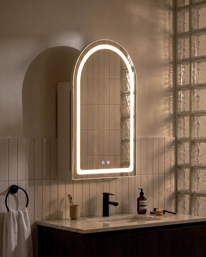 Side perspective of Arcadia arched LED mirror cabinet next to a modern glass brick wall.