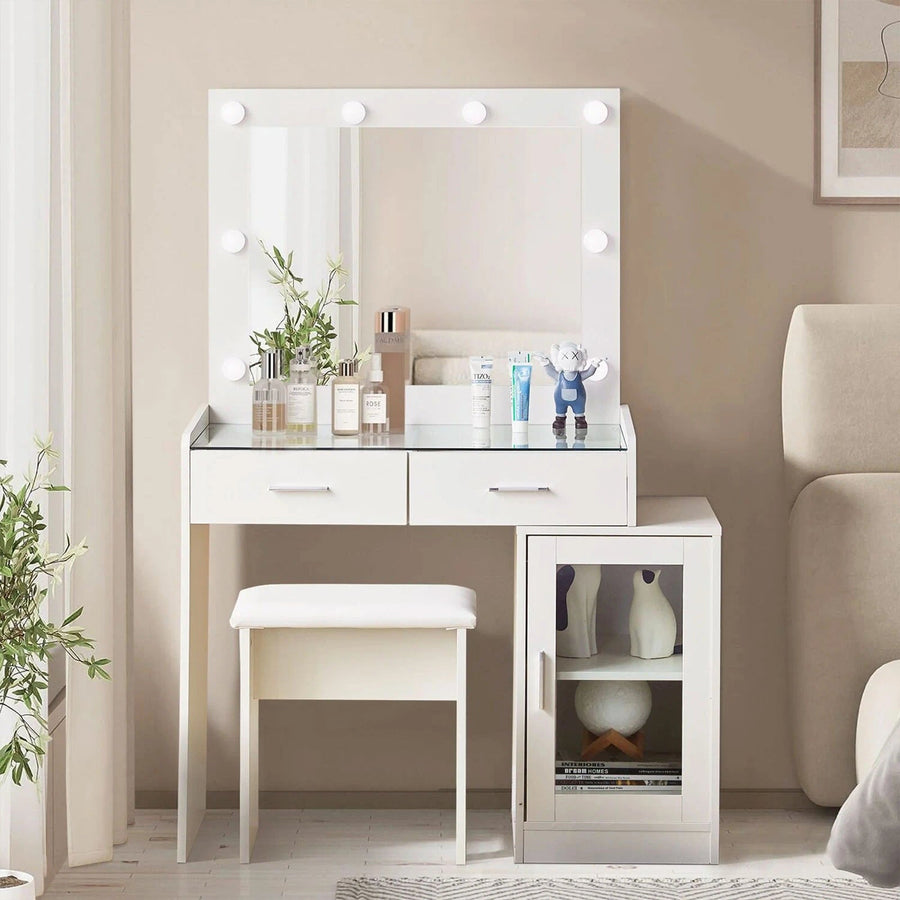 The front view of the white dressing table set, featuring a glass tabletop, a mirror, 8 bulbs, 2 drawers, a cabinet with a glass door, and a stool,  in a bright bedroom.