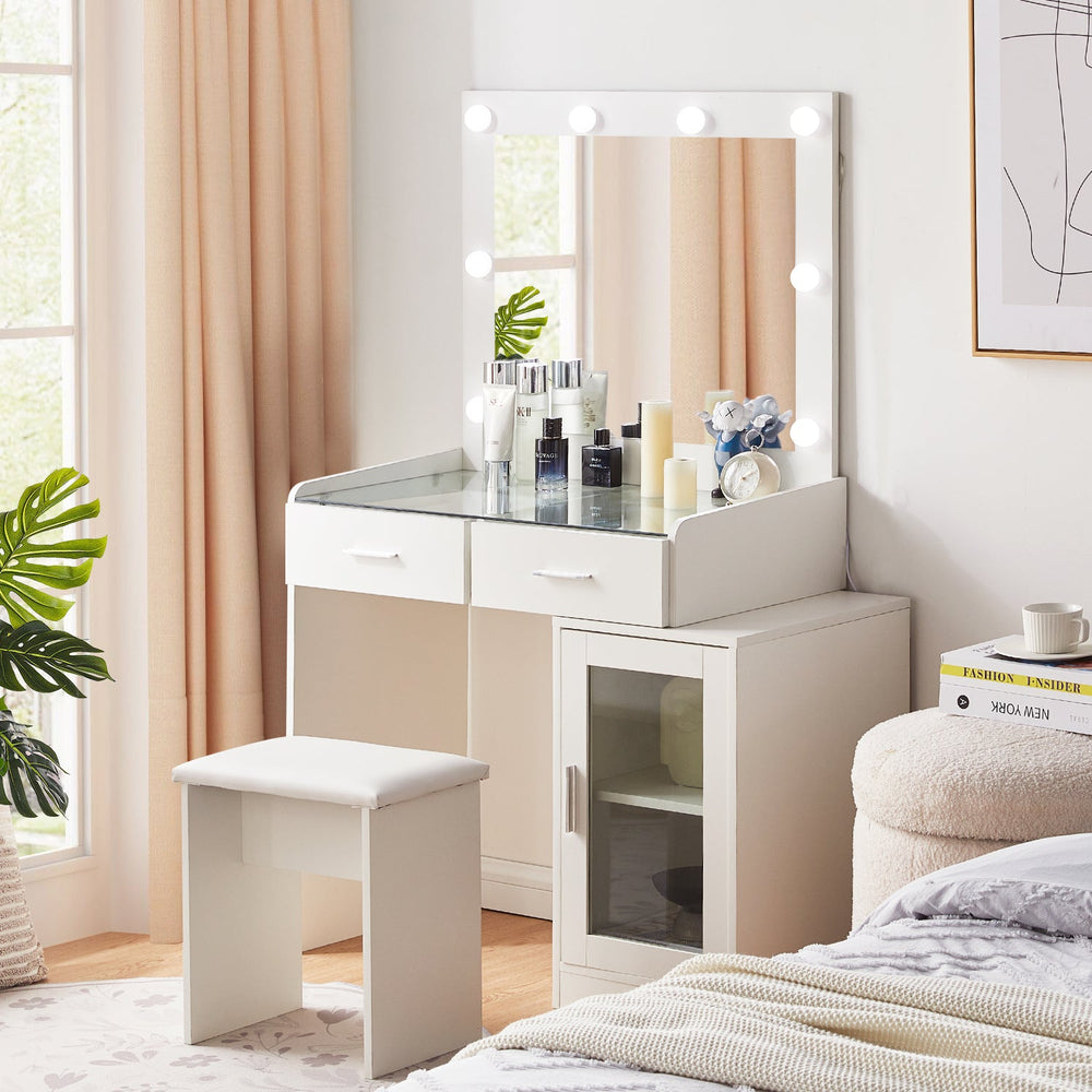 The side view of the white dressing table set, featuring a glass tabletop, a mirror, 8 bulbs, 2 drawers, a cabinet with a glass door, and a stool,  in a bright bedroom.