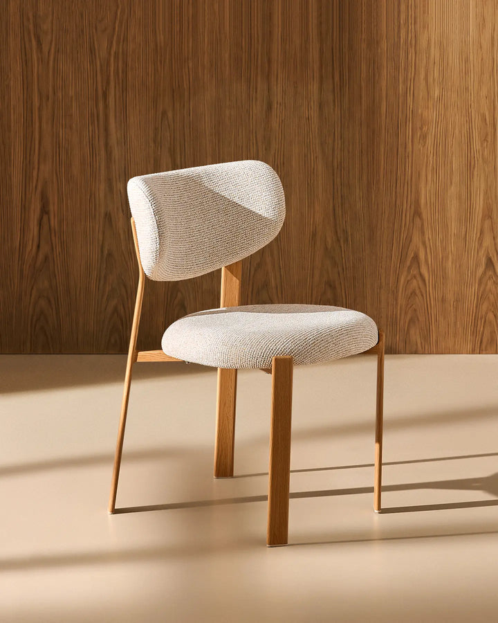 Front view of the Bramley cream chenille dining chair against a warm wood-panelled backdrop, highlighting its sculptural curved backrest