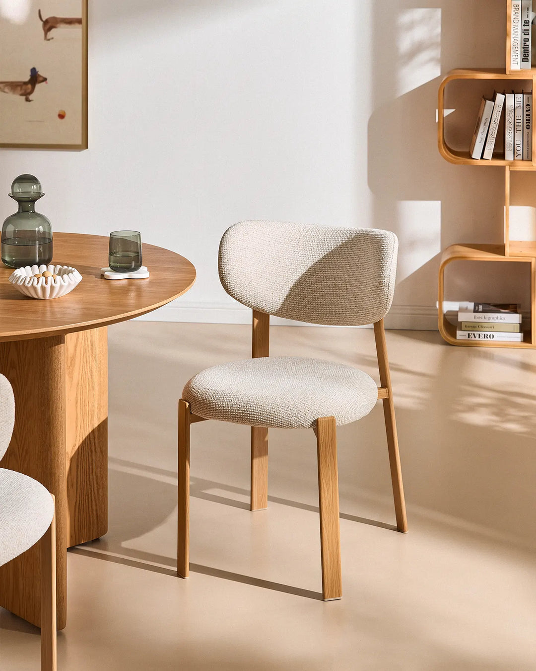 Bramley cream chenille dining chair with curved upholstered backrest and warm wood-effect legs beside a round wooden dining table in a sunlit modern dining room