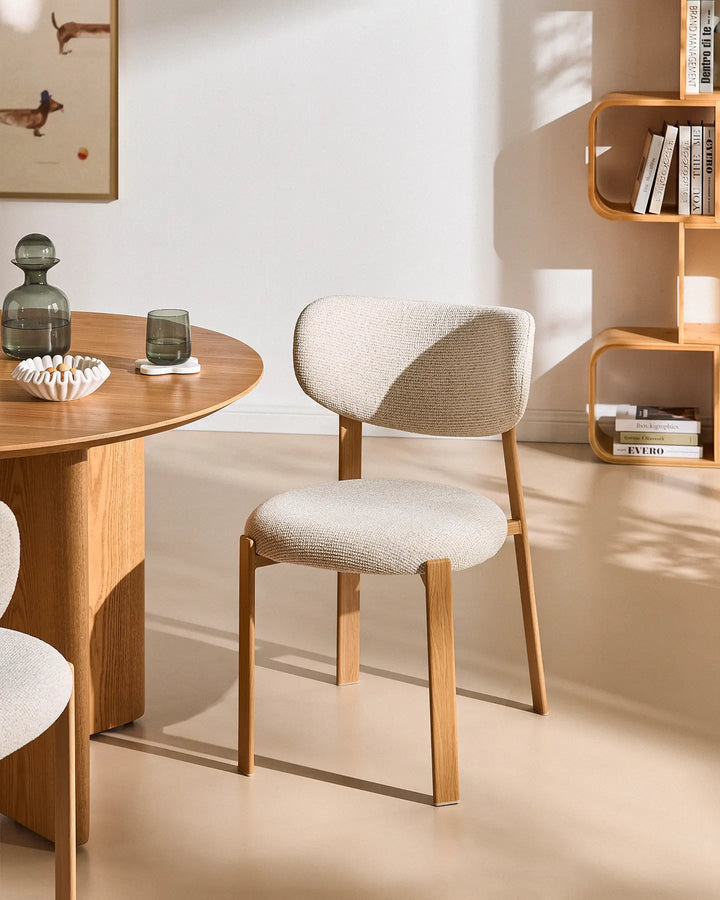 Bramley cream chenille dining chair with curved upholstered backrest and warm wood-effect legs beside a round wooden dining table in a sunlit modern dining room