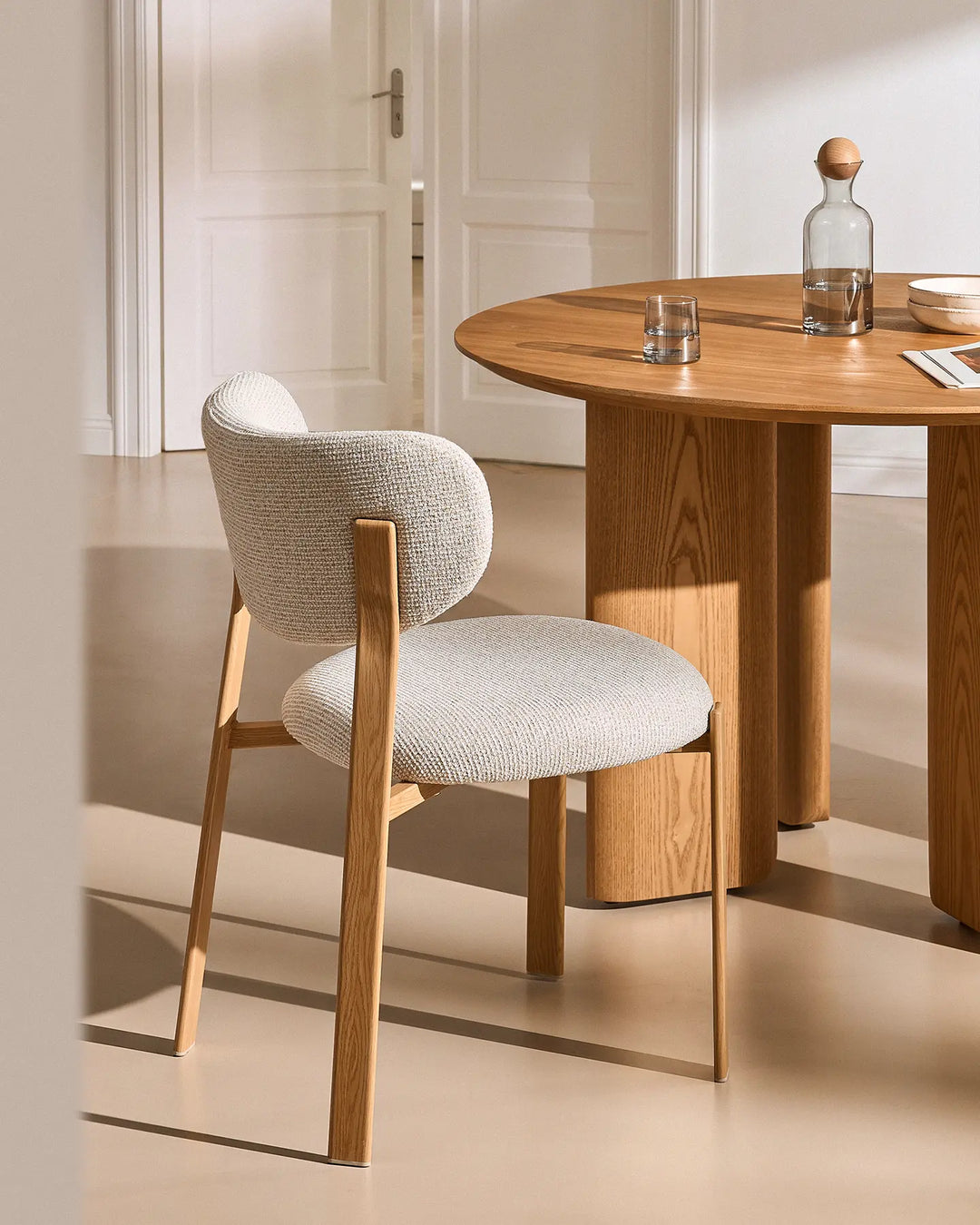 Side view of a Bramley cream chenille dining chair beside a round wooden dining table, showcasing its curved backrest and wooden legs