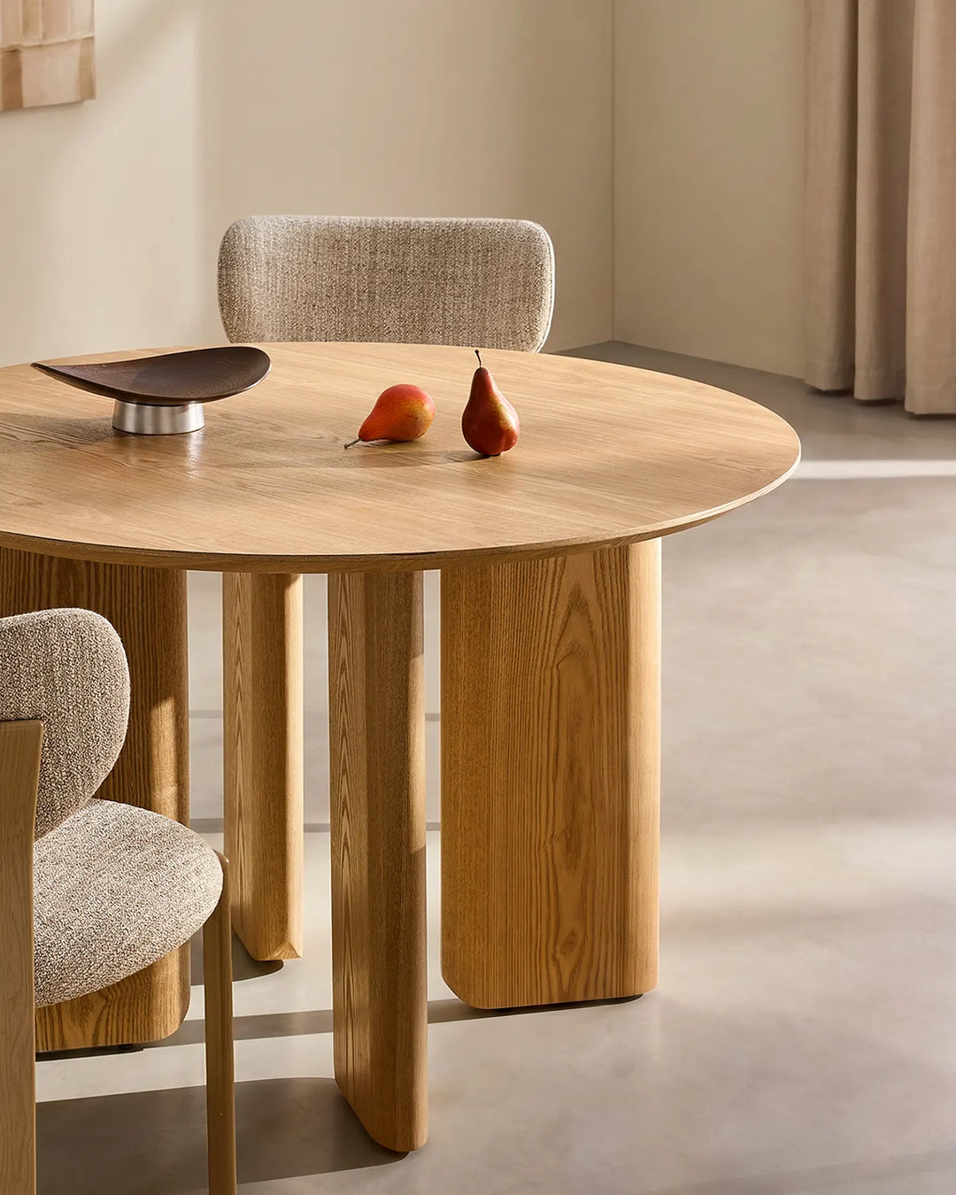 Oak-effect dining table with rounded edges and chunky wooden legs, paired with beige upholstered dining chairs in a warm minimalist dining room. Pears and a raised serving tray decorate the tabletop.