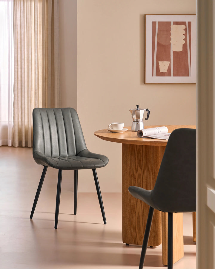 Clair grey faux leather dining chair styled beside a round wooden dining table with black metal legs in a contemporary UK home dining room.