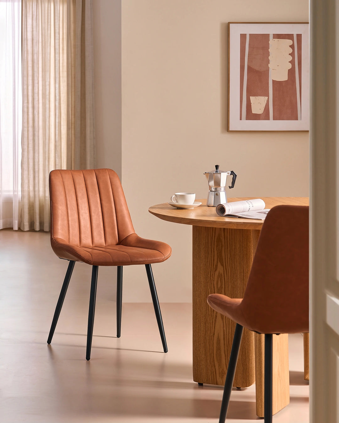 Clair tan brown faux leather dining chair styled beside a round wooden dining table with black metal legs in a contemporary UK home dining room.