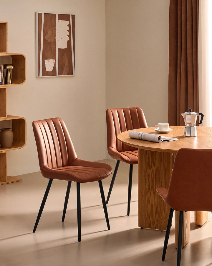 Pair of Clair tan brown faux leather dining chairs arranged around a round timber dining table, showing comfortable seating for social dining.