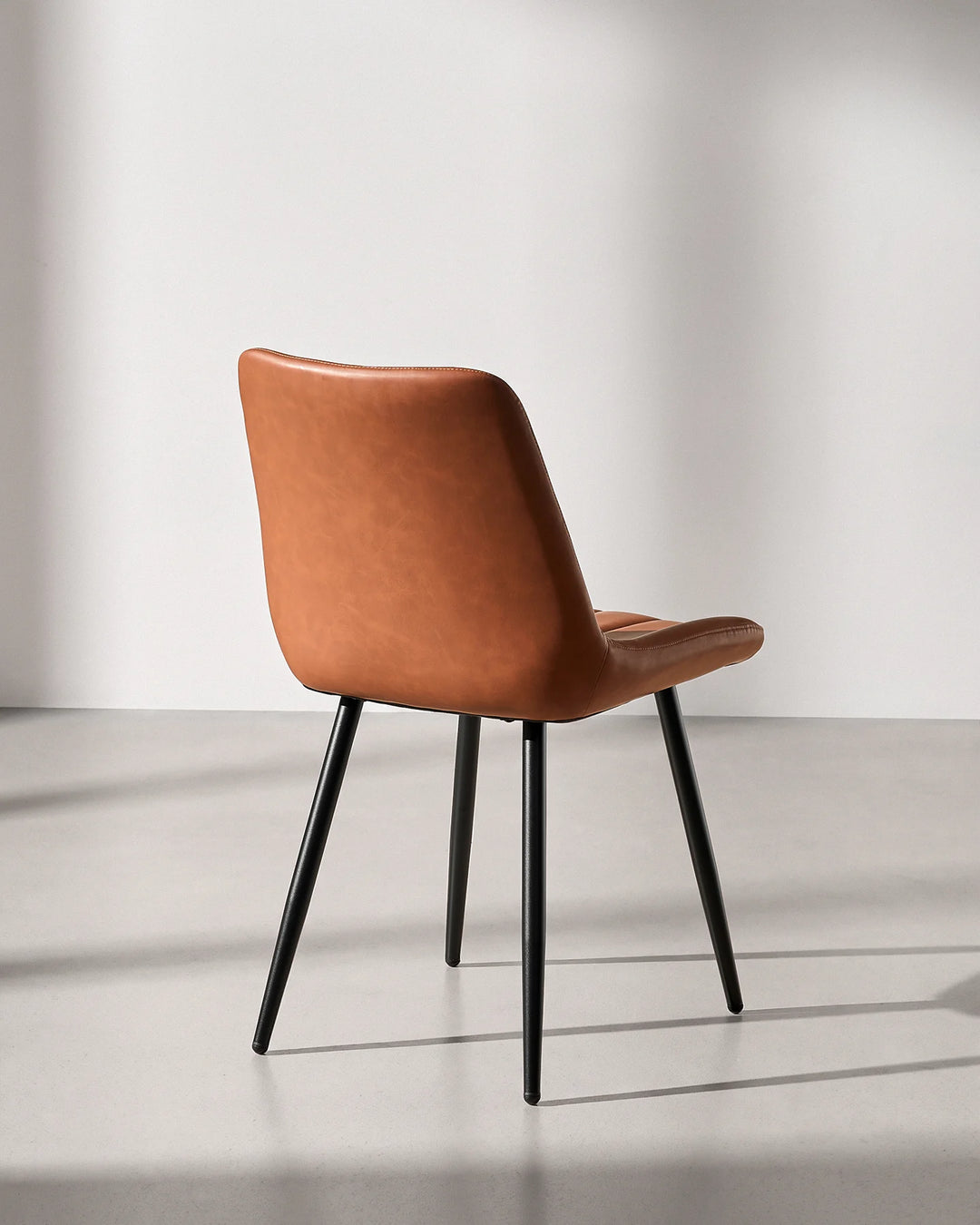 Rear three-quarter view of the Clair tan brown faux leather dining chair, showing smooth wipe-clean upholstery on the back and sturdy steel legs.