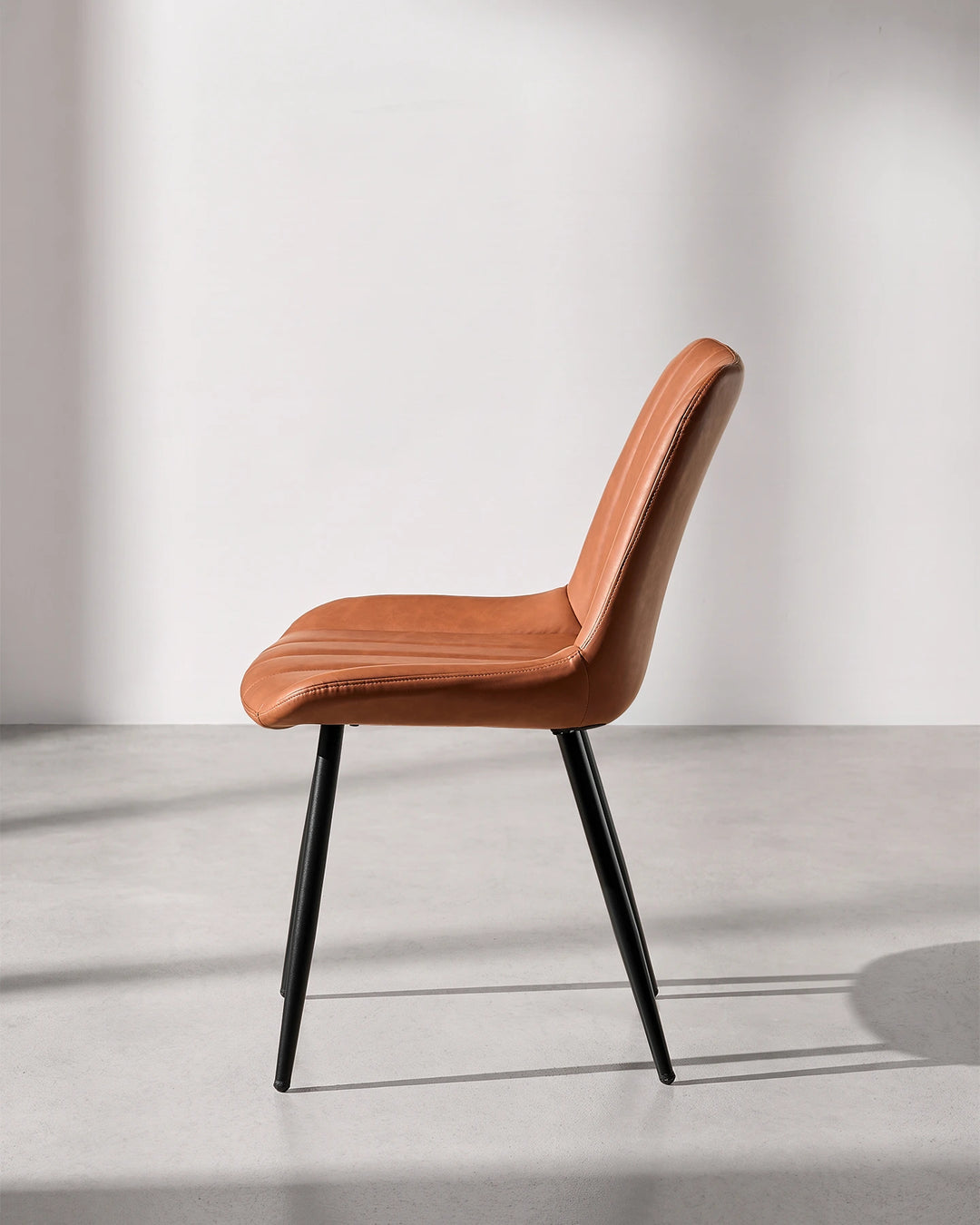 Side profile view of the Clair tan brown faux leather dining chair, displaying the ergonomic curved lumbar back support and slimline silhouette.