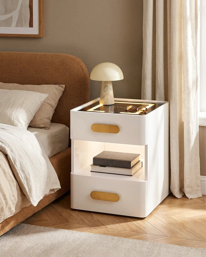 A modern white Clara LED bedside table with gold handles and a glowing shelf, styled in a cozy bedroom next to a bed.