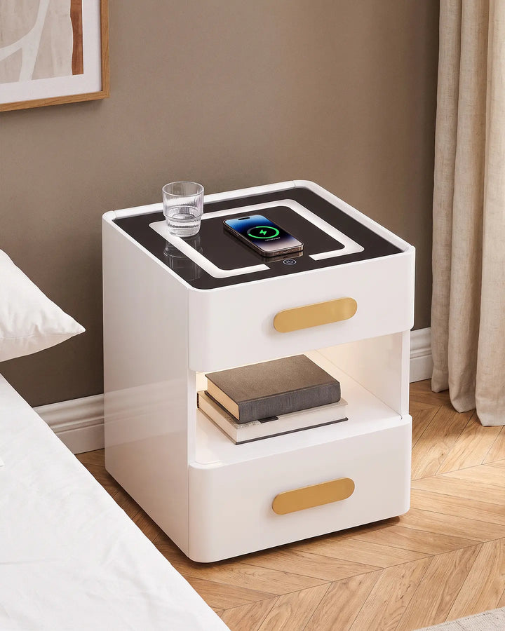 A smartphone charging wirelessly on the glass top of the white Clara LED bedside table, which features a glowing top border, gold handles, and an illuminated shelf with books.