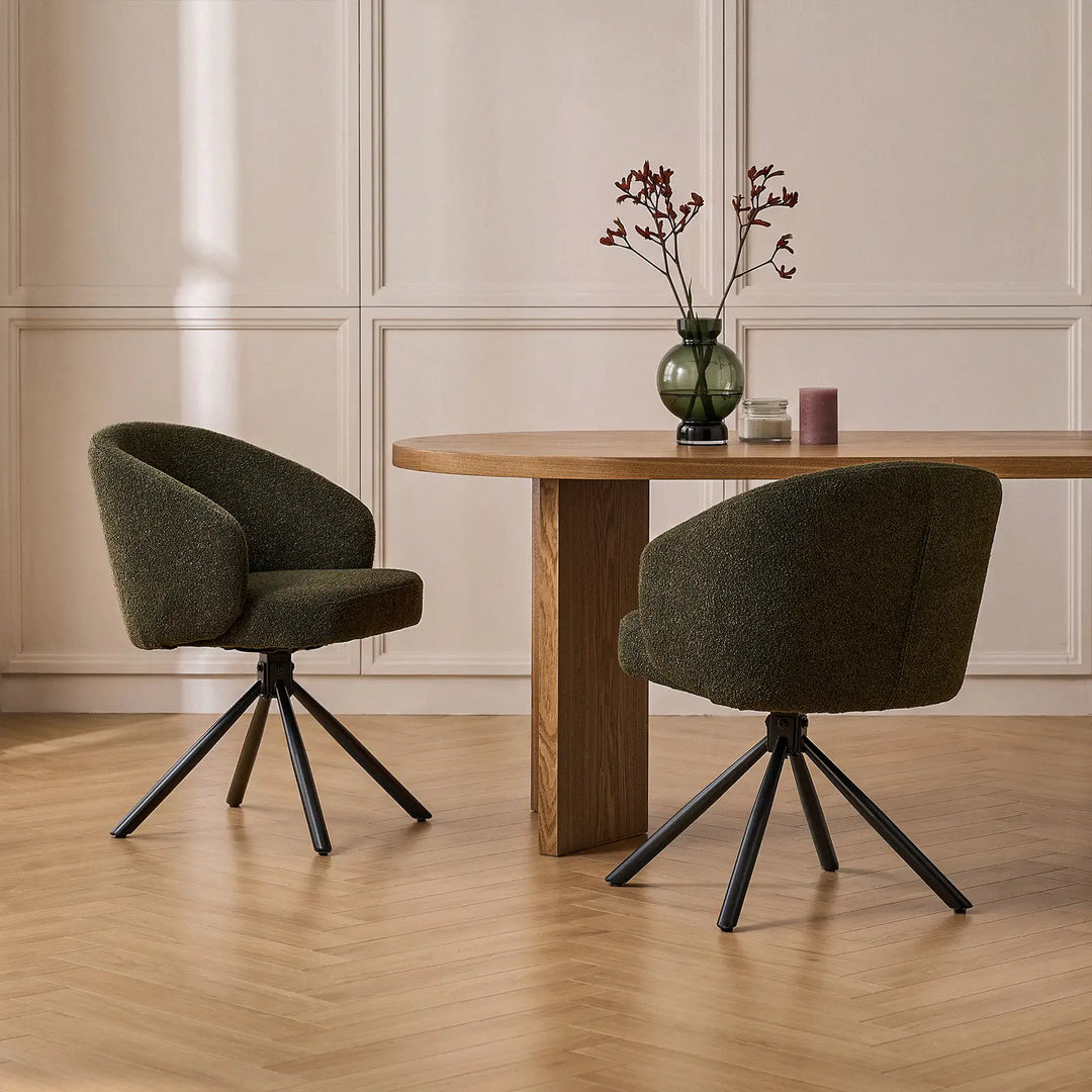 Two Cleo dark green bouclé swivel chairs positioned around an oak dining table with a decorative floral vase in a warm, contemporary dining room interior.