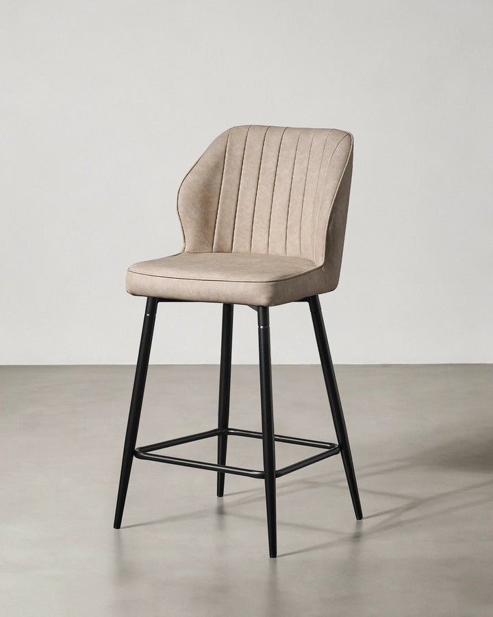 Three-quarter view of a Danforth beige bar stool showing the upholstered seat and backrest with vertical channel stitching and black metal frame