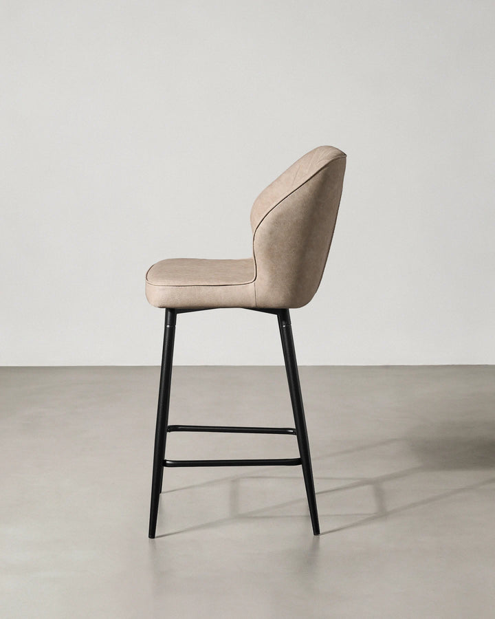 Side profile of a Danforth beige bar stool showing the gently curved upholstered back, cushioned seat and slim black metal legs