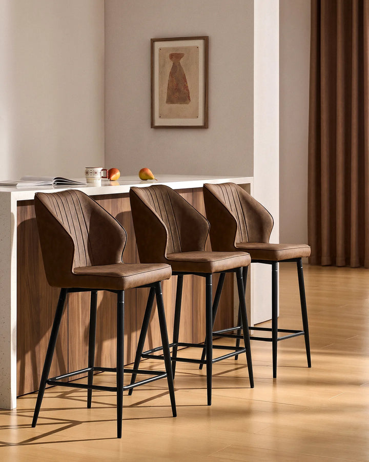 Three Danforth Brown bar stools arranged along a contemporary kitchen island, featuring upholstered seats, vertical stitching and black metal frames