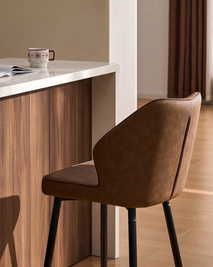 Close-up of a Danforth Brown bar stool showing its softly curved backrest, padded seat, vertical channel stitching and black metal legs