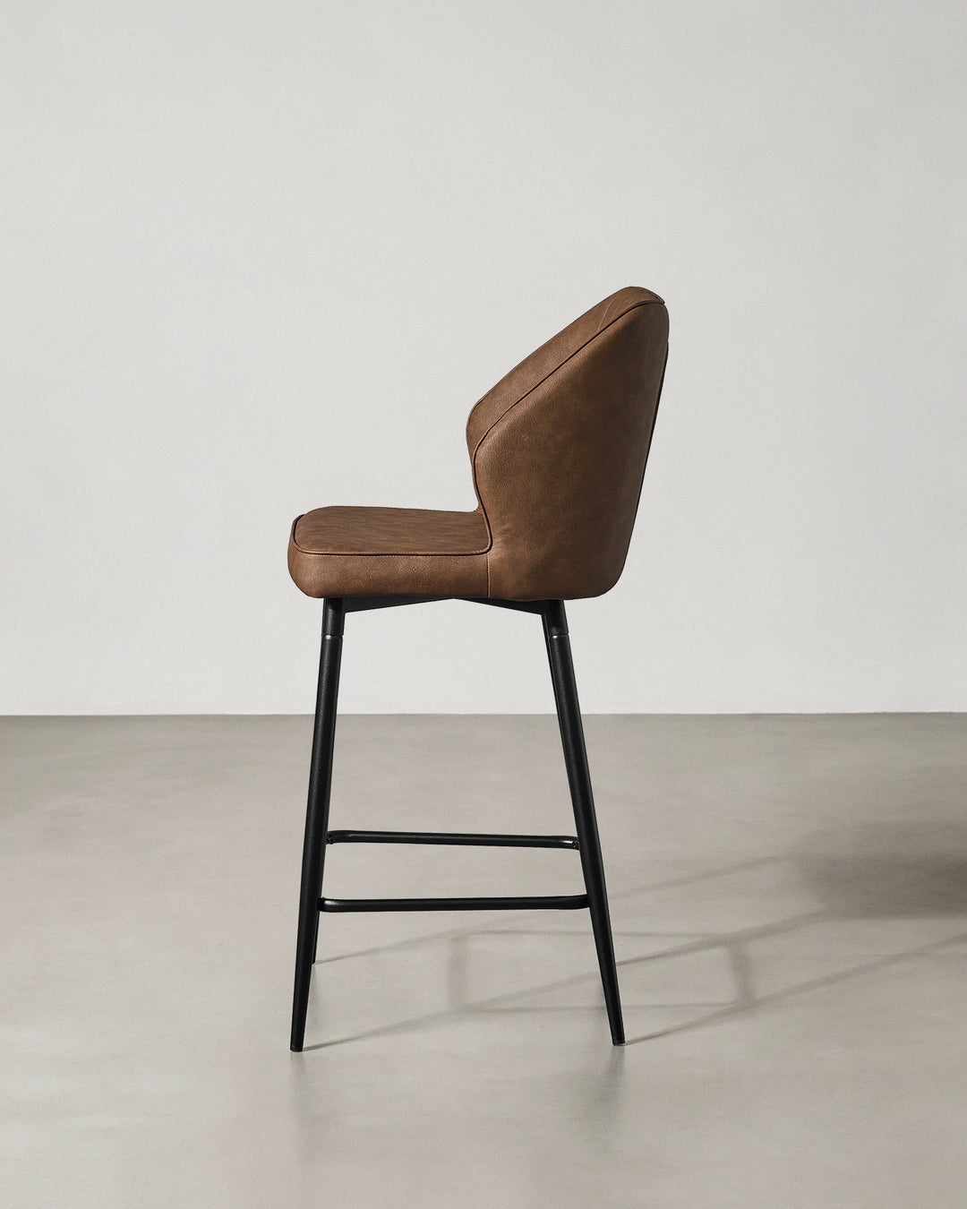 Side profile of a Danforth Brown bar stool showing the gently curved upholstered back, cushioned seat and slim black metal legs