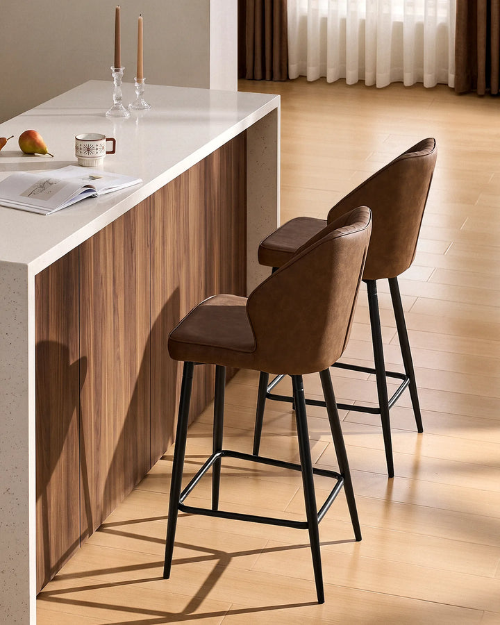 Side view of two Danforth Brown bar stools showing the curved upholstered backrests, slim black metal legs and footrests at a kitchen island