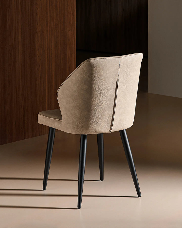 Rear view of the Daviyon beige dining chair, showing its curved upholstered back and slim black metal legs against a warm wood-effect backdrop.