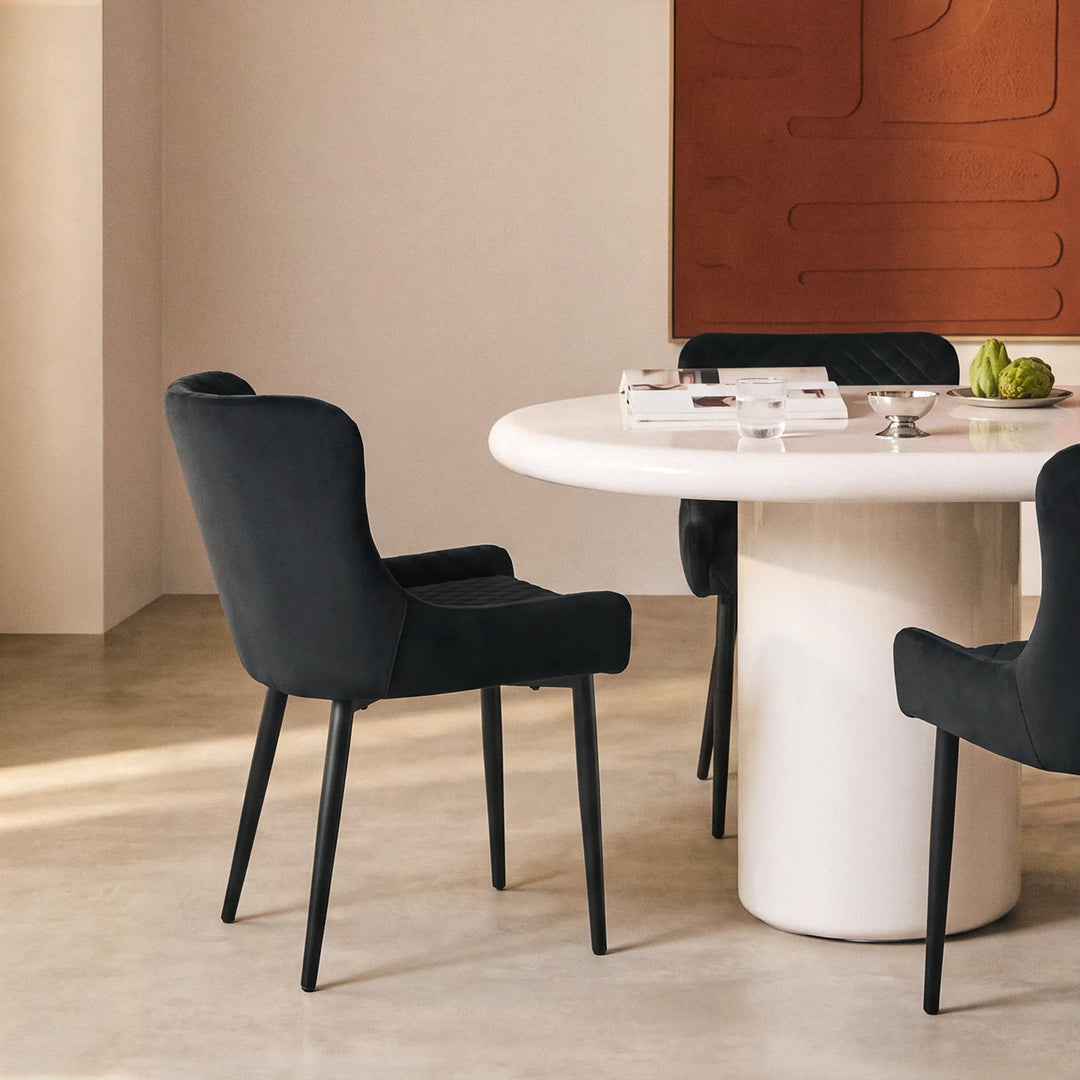 Dundas black velvet dining chair shown from the rear in a modern dining room, paired with a white round dining table and warm wood wall panels.