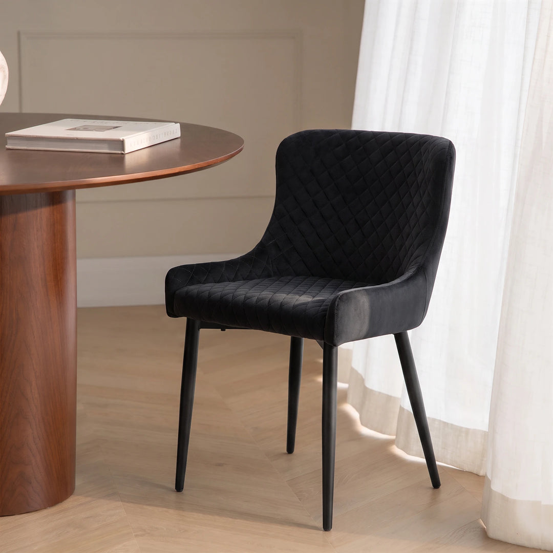 Dundas black velvet dining chair in a contemporary dining space with a white round table, warm wood wall detailing and slim black metal legs.