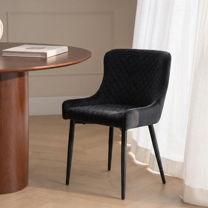Dundas black velvet dining chair in a contemporary dining space with a white round table, warm wood wall detailing and slim black metal legs.
