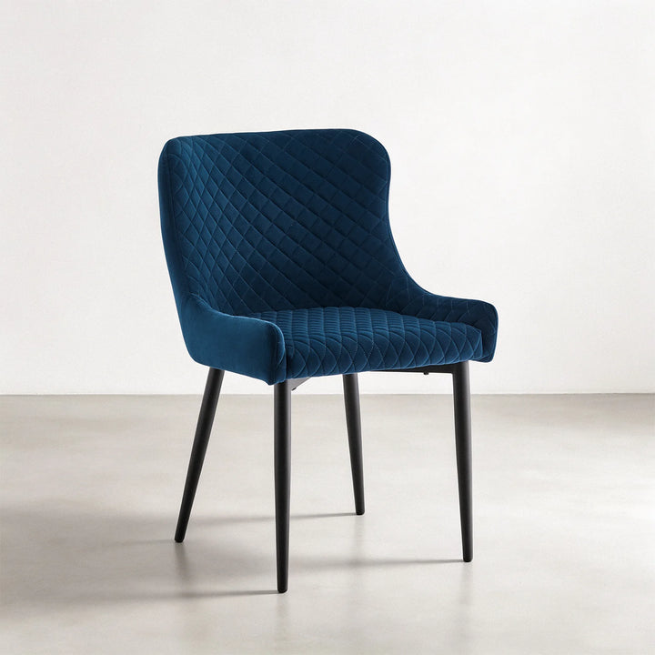 Three-quarter view of a Dundas blue velvet dining chair highlighting the quilted upholstery, supportive armrests and slim black metal frame