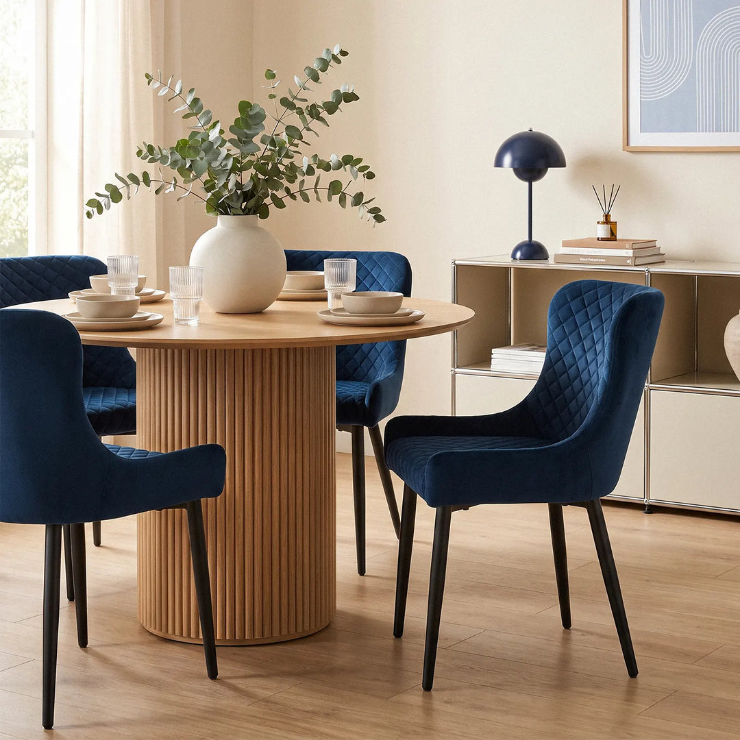 Dundas blue velvet dining chairs with curved armrests and quilted upholstery around a round wooden dining table in a warm modern dining room