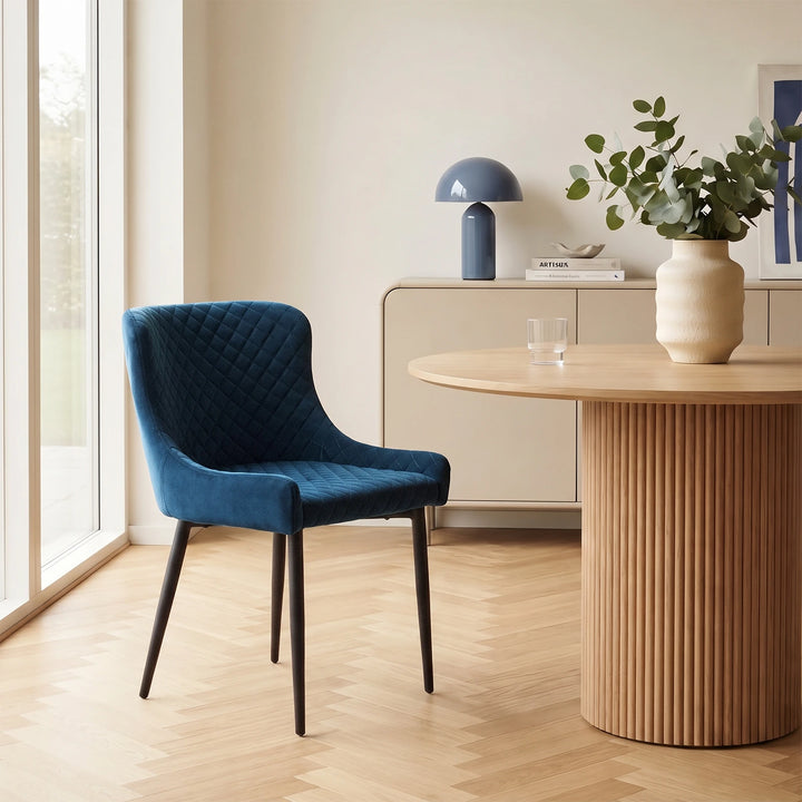 A Dundas blue velvet dining chair with curved backs and black metal legs beside a round wooden dining table