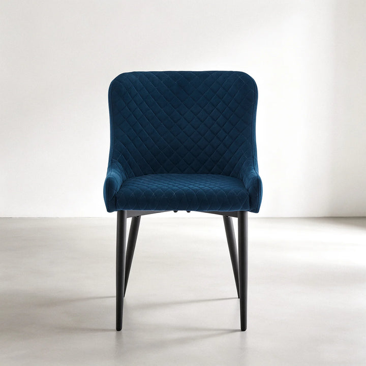 Front view of a Dundas blue velvet dining chair with a gently curved back, padded armrests, quilted seat and black metal legs
