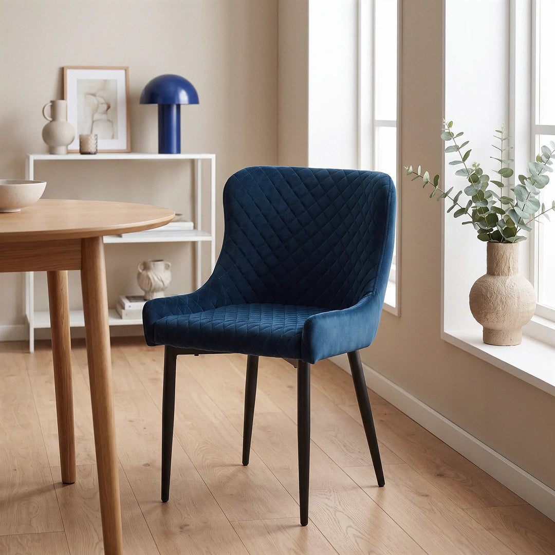 A Dundas blue velvet armchair styled in a modern dining space, featuring quilted upholstery, curved backs and slim black legs