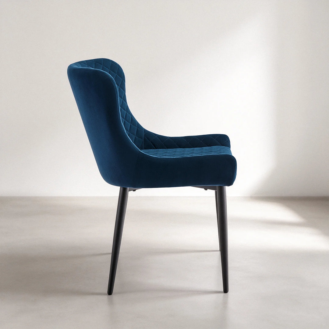 Side view of a Dundas blue velvet dining chair showing its curved backrest, padded armrest, upholstered seat and angled black metal legs