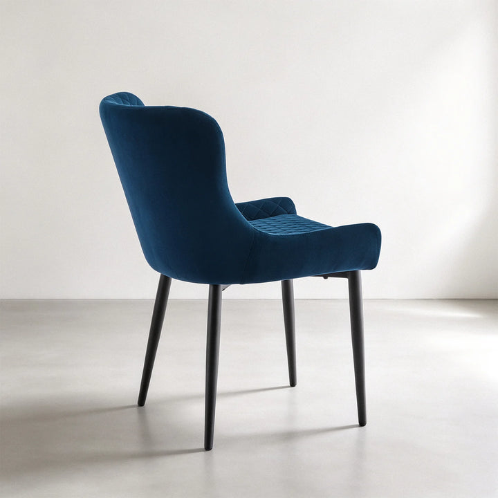 Rear three-quarter view of a Dundas blue velvet dining chair showing the smooth curved back, soft velvet upholstery and black metal legs