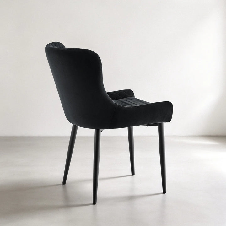 Angled side view of a Dundas black velvet upholstered dining chair with a gently curved back, quilted seat and slim splayed metal legs.