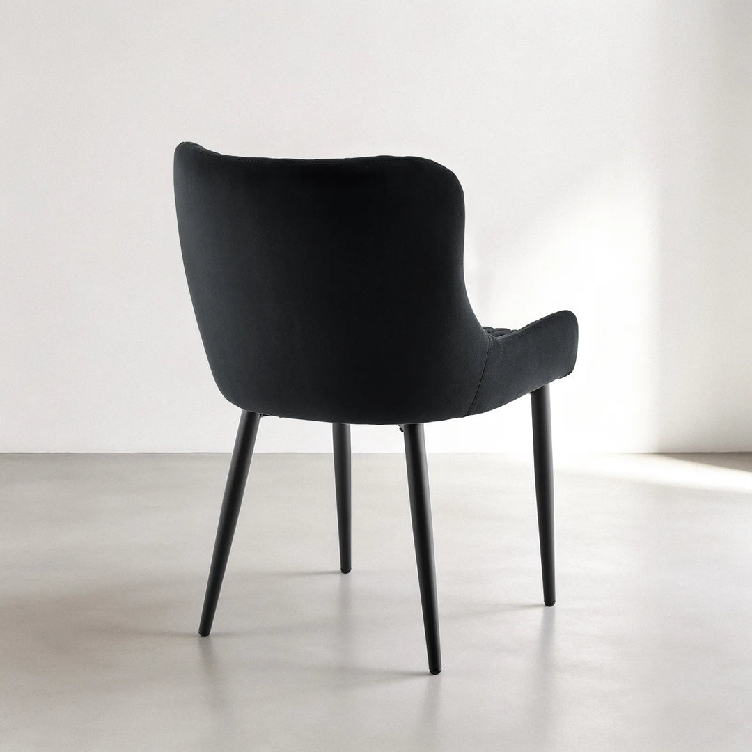 Rear three-quarter view of a Dundas black velvet dining chair showing its smooth curved backrest, padded armrests and black metal leg frame.