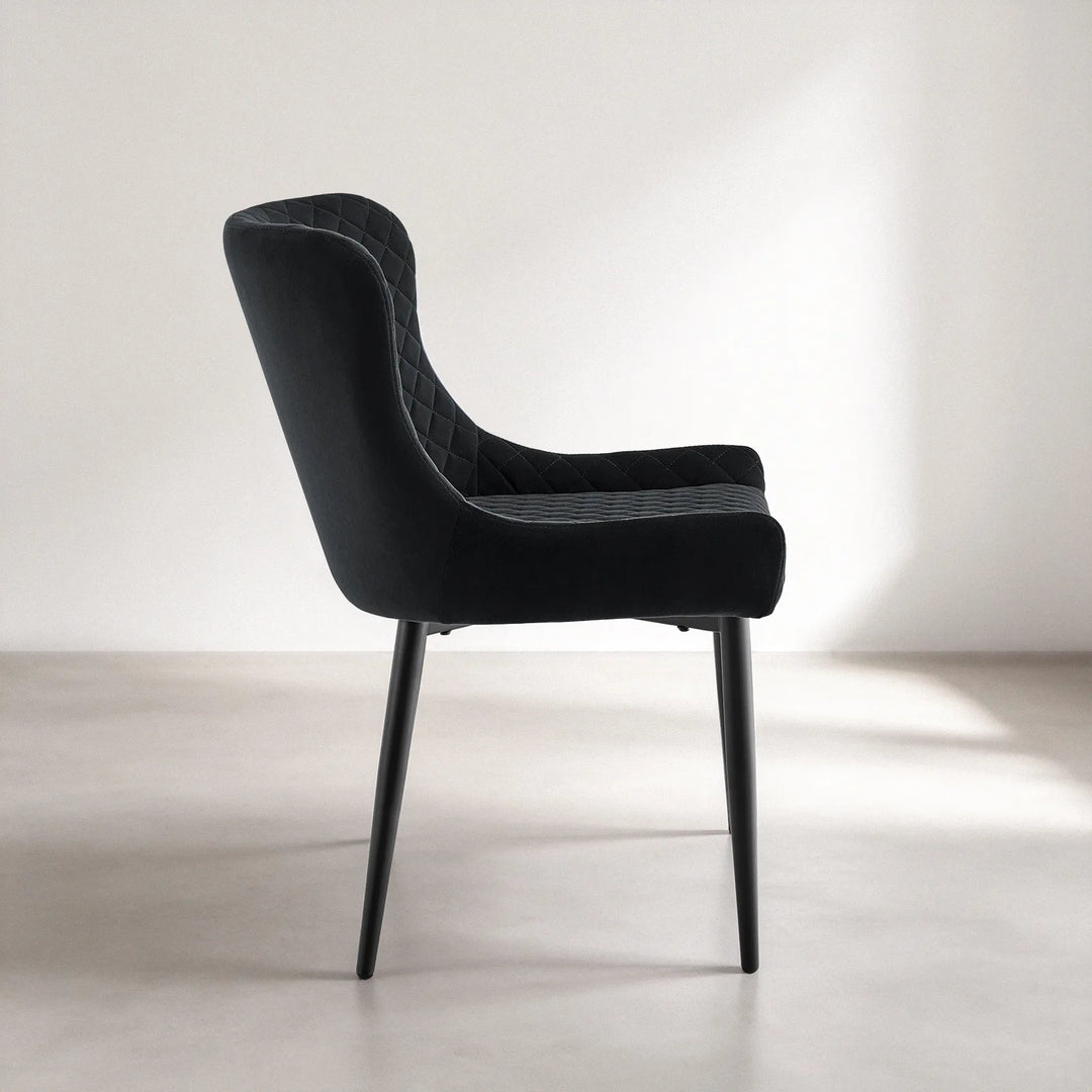 Side profile of a Dundas black velvet dining chair highlighting its curved backrest, padded seat, supportive armrest and angled black metal legs.