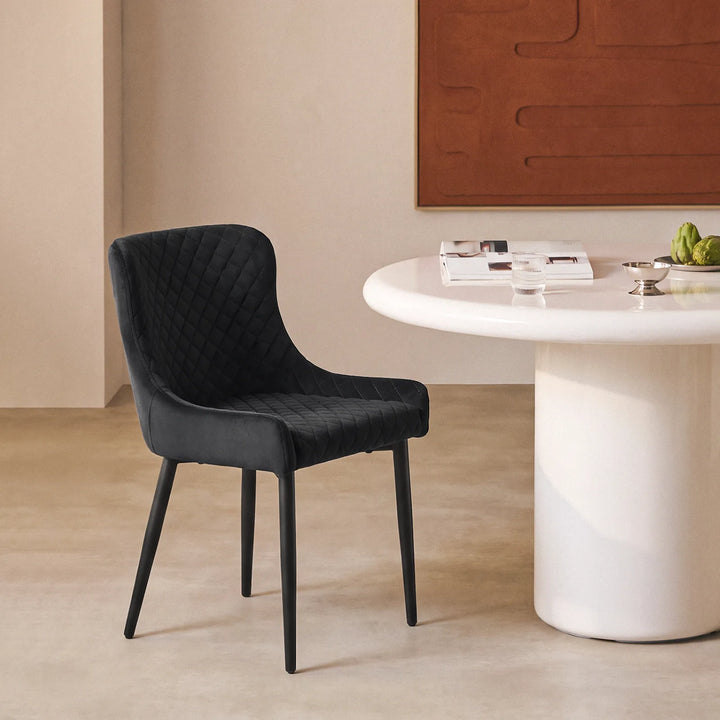 Side view of a Dundas black velvet dining chair beside a modern white dining table, highlighting its curved upholstered back, quilted seat and angled black legs.